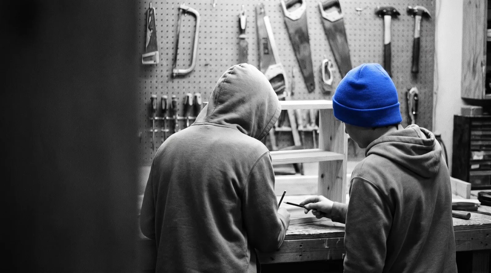Dwóch nastolatków pracuje nad drewnianą półką w warsztacie, jeden ma niebieską czapkę