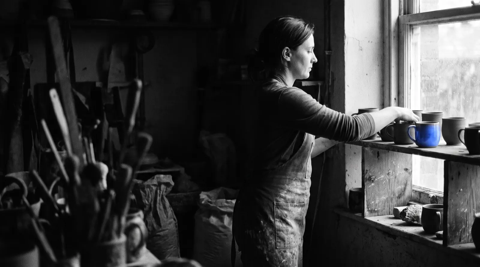 Ceramiczka ustawia kubki na półce w pracowni, jeden z nich jest niebieski