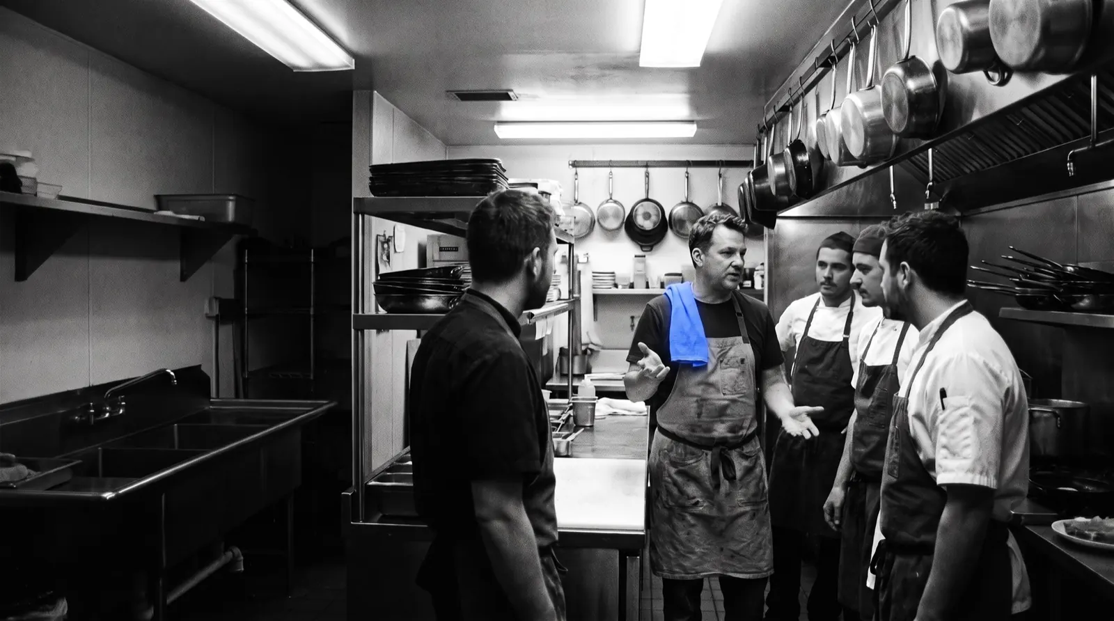 Szef kuchni rozmawia z zespołem w restauracyjnej kuchni, na ramieniu ma niebieską ścierkę